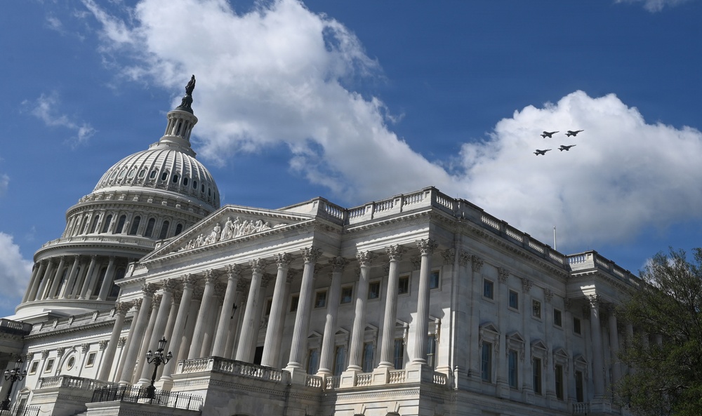 The image shows the United States Capitol Building in Washington, D.C., photographed from a low angle against a partly cloudy blue sky. Four military jet aircraft are visible flying in formation over the building's right side. The neoclassical architecture, including the iconic dome topped with the Statue of Freedom, is prominently featured.