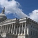 The image shows the United States Capitol Building in Washington, D.C., photographed from a low angle against a partly cloudy blue sky. Four military jet aircraft are visible flying in formation over the building's right side. The neoclassical architecture, including the iconic dome topped with the Statue of Freedom, is prominently featured.