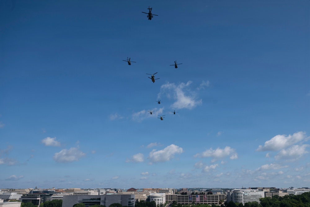 A formation of military helicopters flies over an urban skyline under a mostly clear blue sky with scattered white clouds. The city below appears to be Washington D.C., identifiable by its low-rise federal building architecture and a visible American flag on one of the structures. The helicopters appear to be performing a ceremonial or training flyover in a loose diagonal formation.