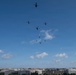 A formation of military helicopters flies over an urban skyline under a mostly clear blue sky with scattered white clouds. The city below appears to be Washington D.C., identifiable by its low-rise federal building architecture and a visible American flag on one of the structures. The helicopters appear to be performing a ceremonial or training flyover in a loose diagonal formation.