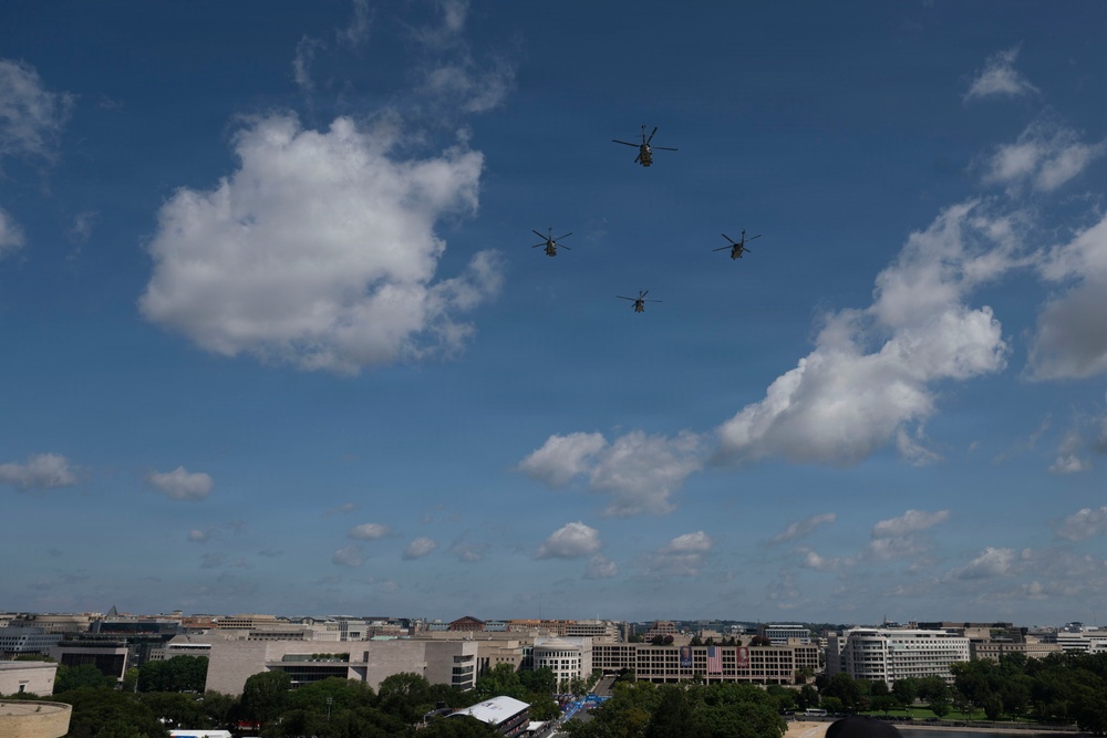Four military helicopters fly in a loose diamond formation over what appears to be Washington D.C., identifiable by the neoclassical and federal-style government buildings visible on the horizon. The aircraft appear to be large tandem-rotor helicopters, consistent with CH-47 Chinook type. The scene suggests a military flyover or ceremonial aerial demonstration.