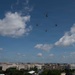 Four military helicopters fly in a loose diamond formation over what appears to be Washington D.C., identifiable by the neoclassical and federal-style government buildings visible on the horizon. The aircraft appear to be large tandem-rotor helicopters, consistent with CH-47 Chinook type. The scene suggests a military flyover or ceremonial aerial demonstration.
