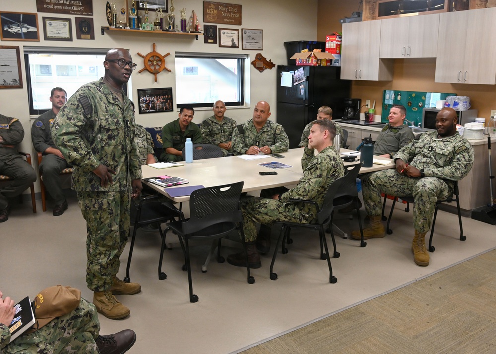 A group of U.S. Navy personnel in Navy Working Uniform (NWU) camouflage are gathered in what appears to be a Chief Petty Officers' mess or meeting room. One individual is standing and addressing the seated group around a conference table. The room is decorated with Navy memorabilia, plaques, trophies, and a sign referencing 'Navy Chief Navy Pride.'