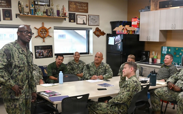 A group of U.S. Navy personnel in Navy Working Uniform (NWU) camouflage are gathered in what appears to be a Chief Petty Officers' mess or meeting room. One individual is standing and addressing the seated group around a conference table. The room is decorated with Navy memorabilia, plaques, trophies, and a sign referencing 'Navy Chief Navy Pride.'