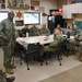 A group of U.S. Navy personnel in Navy Working Uniform (NWU) camouflage are gathered in what appears to be a Chief Petty Officers' mess or meeting room. One individual is standing and addressing the seated group around a conference table. The room is decorated with Navy memorabilia, plaques, trophies, and a sign referencing 'Navy Chief Navy Pride.'