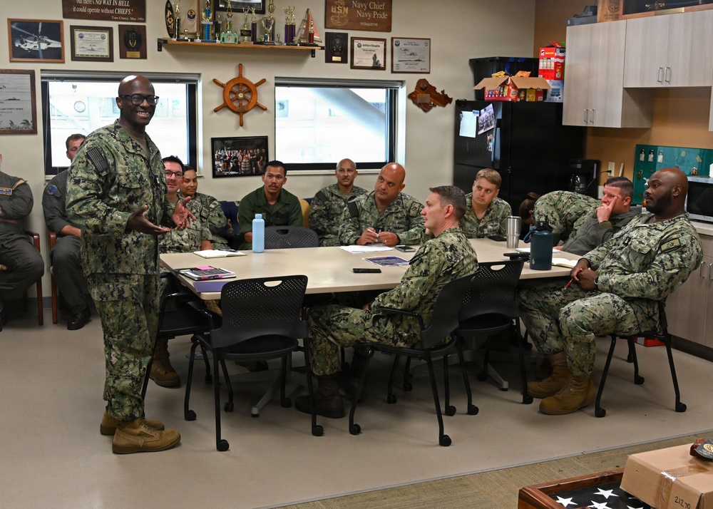 A U.S. Navy briefing or meeting is taking place in what appears to be a Chief Petty Officers' mess or meeting room. A senior enlisted sailor in Navy Working Uniform (NWU Type III camouflage) is standing and addressing a group of approximately 10-12 sailors seated around a conference table. The room is decorated with Navy memorabilia, trophies, plaques, and a ship's wheel, indicating a Chief's quarters or command space.