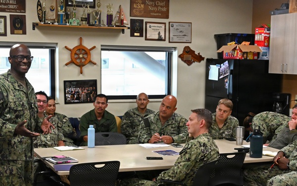 A U.S. Navy briefing or meeting is taking place in what appears to be a Chief Petty Officers' mess or meeting room. A senior enlisted sailor in Navy Working Uniform (NWU Type III camouflage) is standing and addressing a group of approximately 10-12 sailors seated around a conference table. The room is decorated with Navy memorabilia, trophies, plaques, and a ship's wheel, indicating a Chief's quarters or command space.