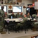 A U.S. Navy briefing or meeting is taking place in what appears to be a Chief Petty Officers' mess or meeting room. A senior enlisted sailor in Navy Working Uniform (NWU Type III camouflage) is standing and addressing a group of approximately 10-12 sailors seated around a conference table. The room is decorated with Navy memorabilia, trophies, plaques, and a ship's wheel, indicating a Chief's quarters or command space.