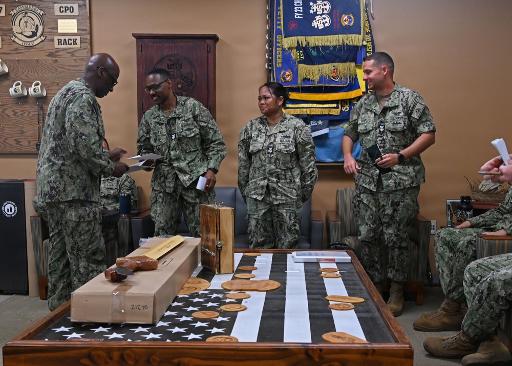 A group of U.S. Navy personnel in Navy Working Uniform (NWU Type III camouflage) are gathered in what appears to be a Chief Petty Officer (CPO) mess or meeting room. A senior enlisted sailor appears to be presenting documents or awards to standing personnel, with a table displaying an American flag shadow box and laser-engraved wooden items in the foreground. The setting suggests a formal recognition or awards ceremony.