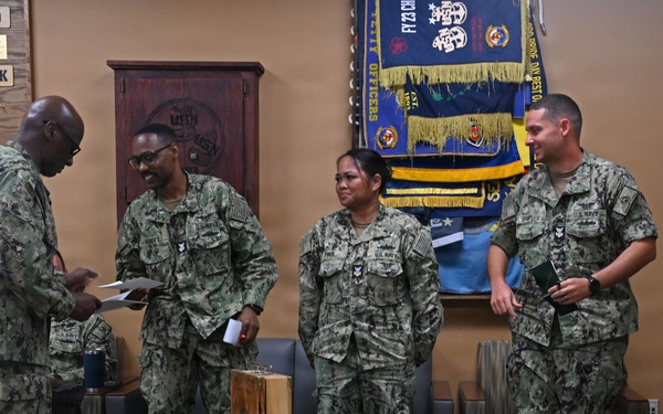 A group of U.S. Navy personnel in Navy Working Uniform (NWU Type III camouflage) are gathered in what appears to be a Chief Petty Officer (CPO) mess or meeting room. A senior enlisted sailor appears to be presenting documents or awards to standing personnel, with a table displaying an American flag shadow box and laser-engraved wooden items in the foreground. The setting suggests a formal recognition or awards ceremony.