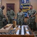 A group of U.S. Navy personnel in Navy Working Uniform (NWU Type III camouflage) are gathered in what appears to be a Chief Petty Officer (CPO) mess or meeting room. A senior enlisted sailor appears to be presenting documents or awards to standing personnel, with a table displaying an American flag shadow box and laser-engraved wooden items in the foreground. The setting suggests a formal recognition or awards ceremony.