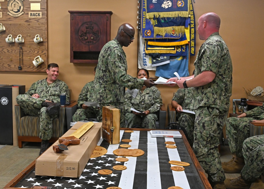 A group of U.S. Navy personnel in NWU (Navy Working Uniform) camouflage are gathered in what appears to be a command or CPO (Chief Petty Officer) mess room. Two sailors in the foreground are exchanging documents, while others are seated around the room. A table in the foreground displays an American flag (black and white), multiple laser-engraved wooden medallions/coins, and packaged items.
