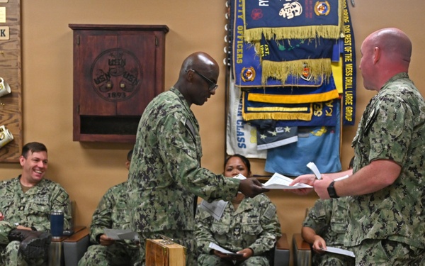 A group of U.S. Navy personnel in NWU (Navy Working Uniform) camouflage are gathered in what appears to be a command or CPO (Chief Petty Officer) mess room. Two sailors in the foreground are exchanging documents, while others are seated around the room. A table in the foreground displays an American flag (black and white), multiple laser-engraved wooden medallions/coins, and packaged items.