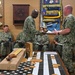 A group of U.S. Navy personnel in NWU (Navy Working Uniform) camouflage are gathered in what appears to be a command or CPO (Chief Petty Officer) mess room. Two sailors in the foreground are exchanging documents, while others are seated around the room. A table in the foreground displays an American flag (black and white), multiple laser-engraved wooden medallions/coins, and packaged items.