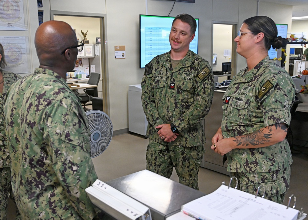 Three U.S. Navy personnel in NWU Type III (woodland camouflage) uniforms are engaged in conversation in what appears to be a military administrative office environment. The central figure, whose name tape reads 'PENFIELD,' is smiling while speaking with a colleague whose back is to the camera. A female sailor stands to the right, also participating in the discussion.
