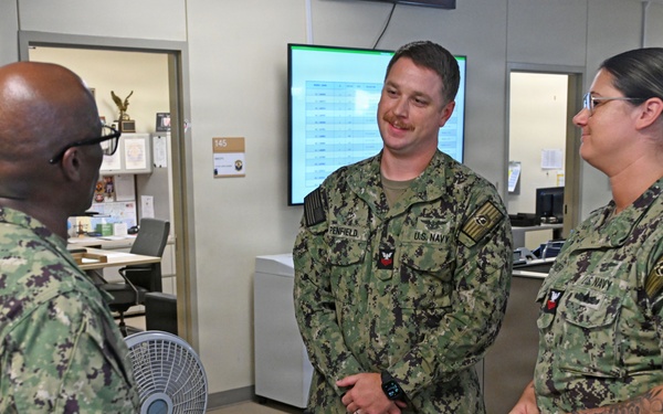 Three U.S. Navy personnel in NWU Type III (woodland camouflage) uniforms are engaged in conversation in what appears to be a military administrative office environment. The central figure, whose name tape reads 'PENFIELD,' is smiling while speaking with a colleague whose back is to the camera. A female sailor stands to the right, also participating in the discussion.
