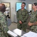 Three U.S. Navy personnel in NWU Type III (woodland camouflage) uniforms are engaged in conversation in what appears to be a military administrative office environment. The central figure, whose name tape reads 'PENFIELD,' is smiling while speaking with a colleague whose back is to the camera. A female sailor stands to the right, also participating in the discussion.