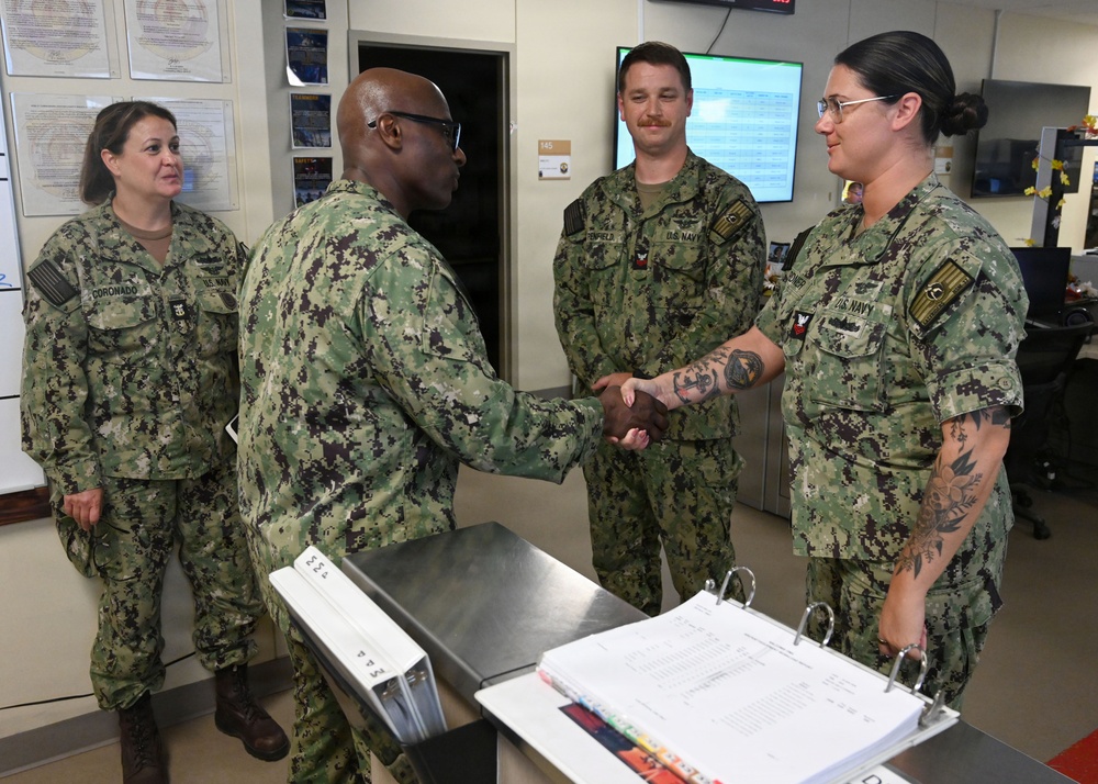 Four U.S. Navy personnel in NWU Type III (green camouflage) uniforms are gathered in what appears to be a military office or operations center. A senior officer is shaking hands with a female sailor who has visible tattoos on her arms, suggesting a formal recognition or greeting ceremony. Two other sailors, including one identified by nametape as 'Coronado,' observe the interaction.