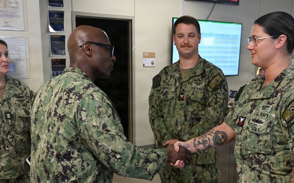 Four U.S. Navy personnel in NWU Type III (green camouflage) uniforms are gathered in what appears to be a military office or operations center. A senior officer is shaking hands with a female sailor who has visible tattoos on her arms, suggesting a formal recognition or greeting ceremony. Two other sailors, including one identified by nametape as 'Coronado,' observe the interaction.