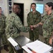 Four U.S. Navy personnel in NWU Type III (green camouflage) uniforms are gathered in what appears to be a military office or operations center. A senior officer is shaking hands with a female sailor who has visible tattoos on her arms, suggesting a formal recognition or greeting ceremony. Two other sailors, including one identified by nametape as 'Coronado,' observe the interaction.