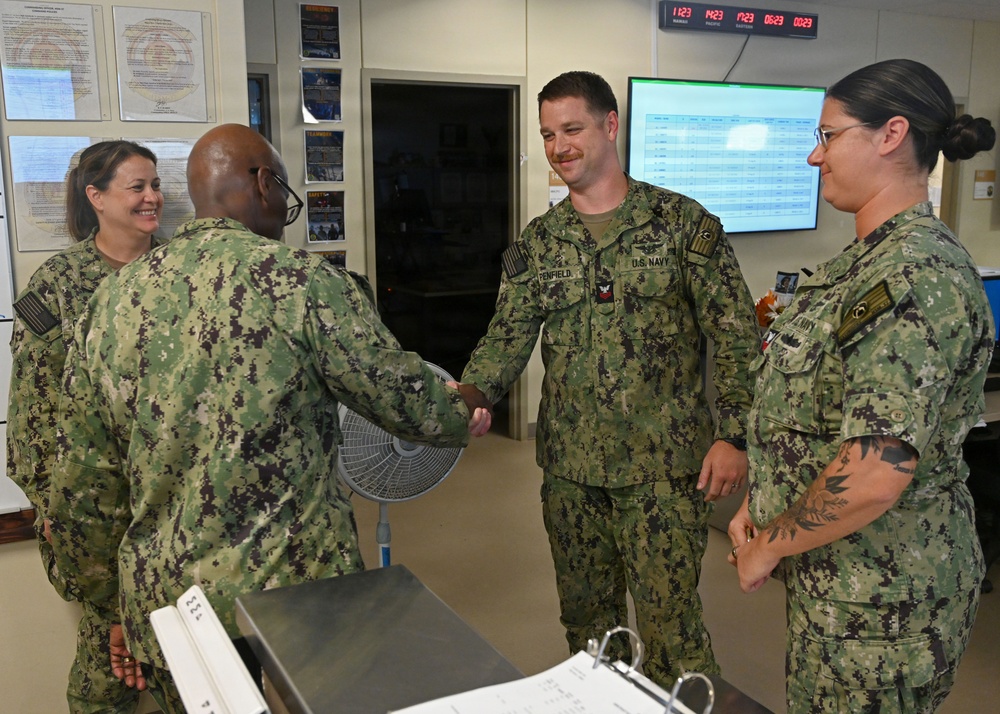 Four U.S. Navy personnel in NWU Type III (woodland digital camouflage) uniforms are gathered in what appears to be a military operations or administrative office. Two individuals are shaking hands in what appears to be a formal recognition or greeting ceremony, with two others observing and smiling. The setting includes a multi-timezone digital clock display, a monitor with data, and bulletin boards with posted documents.