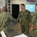 Four U.S. Navy personnel in NWU Type III (woodland digital camouflage) uniforms are gathered in what appears to be a military operations or administrative office. Two individuals are shaking hands in what appears to be a formal recognition or greeting ceremony, with two others observing and smiling. The setting includes a multi-timezone digital clock display, a monitor with data, and bulletin boards with posted documents.