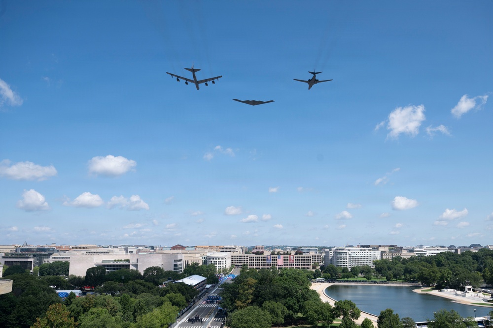 Three military aircraft are conducting a flyover above Washington D.C., identifiable by the National Mall area and surrounding federal buildings. The formation includes what appears to be a B-52 Stratofortress on the left, a B-2 Spirit stealth bomber in the center, and a B-1 Lancer on the right. The scene suggests a ceremonial or commemorative flyover event, with a crowd visible along the Mall below.