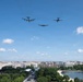 Three military aircraft are conducting a flyover above Washington D.C., identifiable by the National Mall area and surrounding federal buildings. The formation includes what appears to be a B-52 Stratofortress on the left, a B-2 Spirit stealth bomber in the center, and a B-1 Lancer on the right. The scene suggests a ceremonial or commemorative flyover event, with a crowd visible along the Mall below.