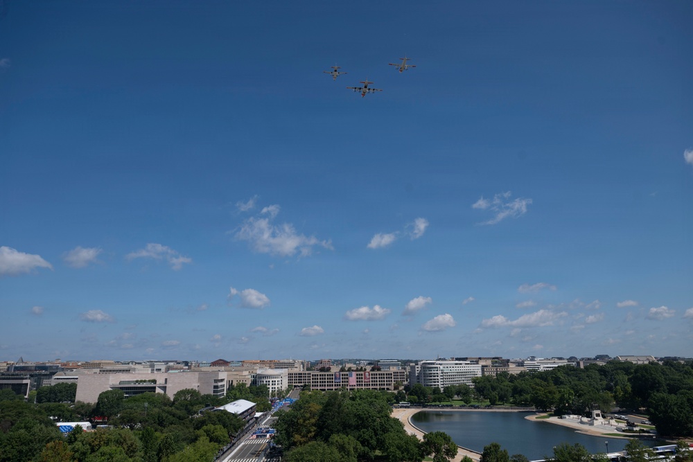 An aerial view of Washington D.C. showing a formation of three military aircraft flying over the National Mall area. The cityscape includes recognizable federal buildings, tree-lined boulevards, and a reflecting pool or water feature visible in the lower right. The scene appears to capture a military flyover event, likely associated with a national celebration or commemoration.