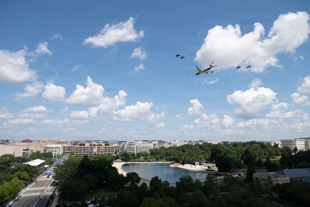 An aerial flyover formation is captured over what appears to be Washington D.C., featuring a large blue-and-white aircraft consistent with Air Force One accompanied by smaller military fighter jets. The scene is photographed from an elevated vantage point showing a urban park landscape with a reflecting pool or small lake below. The image was taken on a partly cloudy summer day with lush green tree canopy visible throughout.
