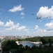An aerial flyover formation is captured over what appears to be Washington D.C., featuring a large blue-and-white aircraft consistent with Air Force One accompanied by smaller military fighter jets. The scene is photographed from an elevated vantage point showing a urban park landscape with a reflecting pool or small lake below. The image was taken on a partly cloudy summer day with lush green tree canopy visible throughout.