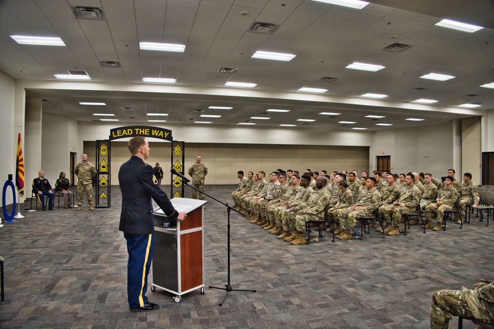 A U.S. Army officer in dress blues is addressing a large formation of soldiers seated in rows at a formal military ceremony. A banner reading 'LEAD THE WAY' with Sergeant Major rank insignia is displayed behind the podium, suggesting this is an NCO-related event. The ceremony appears to take place in a large indoor conference or assembly hall.