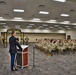 A U.S. Army officer in dress blues is addressing a large formation of soldiers seated in rows at a formal military ceremony. A banner reading 'LEAD THE WAY' with Sergeant Major rank insignia is displayed behind the podium, suggesting this is an NCO-related event. The ceremony appears to take place in a large indoor conference or assembly hall.