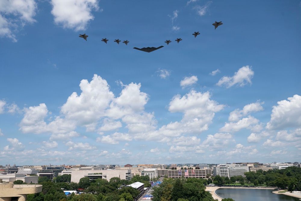 A military flyover formation is captured above what appears to be Washington D.C., featuring a B-2 Spirit stealth bomber as the centerpiece flanked by multiple fighter jets in a V-shaped formation. The urban landscape below includes recognizable federal-style buildings, tree-lined areas, and a body of water consistent with the National Mall area. The image was likely taken during a national celebration or military commemoration event.