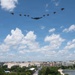 A military flyover formation is captured above what appears to be Washington D.C., featuring a B-2 Spirit stealth bomber as the centerpiece flanked by multiple fighter jets in a V-shaped formation. The urban landscape below includes recognizable federal-style buildings, tree-lined areas, and a body of water consistent with the National Mall area. The image was likely taken during a national celebration or military commemoration event.