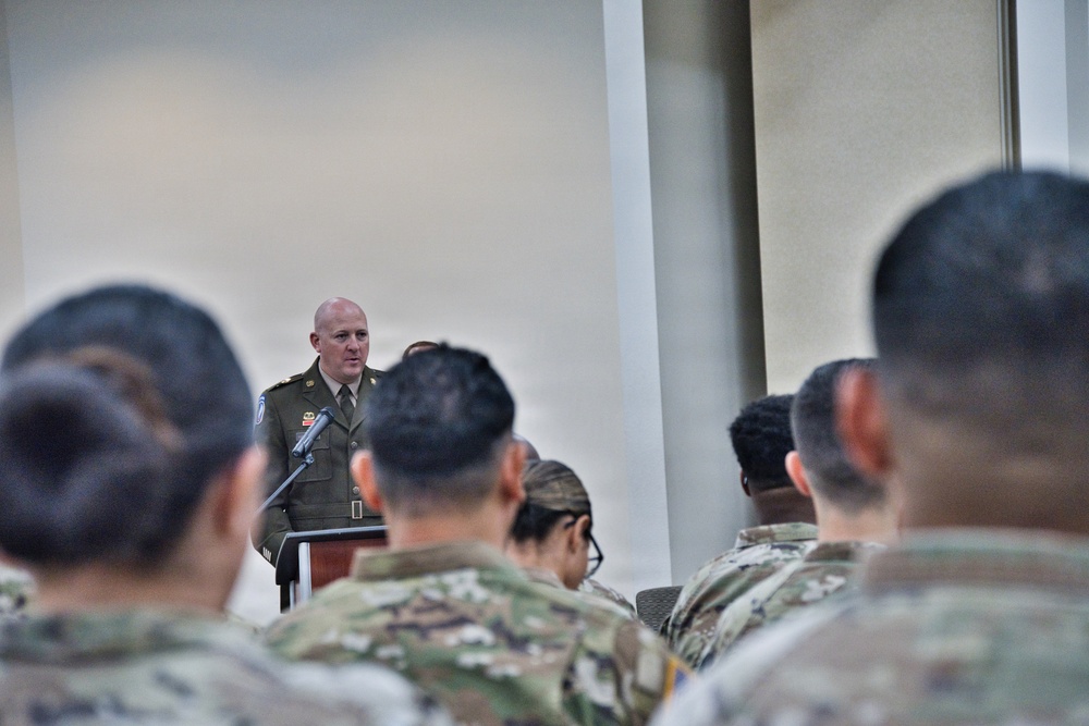 A military officer in dress uniform stands at a podium with a microphone, addressing an audience of soldiers seated in the foreground. The speaker appears to be delivering a formal briefing, ceremony speech, or official address. The audience members are wearing camouflage (OCP/MultiCam) uniforms, suggesting a mixed-formality military event.