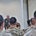 A military officer in dress uniform stands at a podium with a microphone, addressing an audience of soldiers seated in the foreground. The speaker appears to be delivering a formal briefing, ceremony speech, or official address. The audience members are wearing camouflage (OCP/MultiCam) uniforms, suggesting a mixed-formality military event.