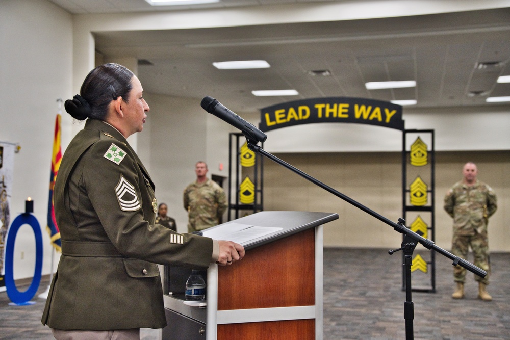 A senior U.S. Army non-commissioned officer, identifiable by Sergeant Major rank insignia, delivers a speech at a podium during a military ceremony. The event appears to be an NCO-related promotion or recognition ceremony, evidenced by the 'LEAD THE WAY' arch decorated with Sergeant Major chevron insignia in the background. Several soldiers in Army Combat Uniforms (ACUs) stand at attention in the background.