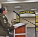 A senior U.S. Army non-commissioned officer, identifiable by Sergeant Major rank insignia, delivers a speech at a podium during a military ceremony. The event appears to be an NCO-related promotion or recognition ceremony, evidenced by the 'LEAD THE WAY' arch decorated with Sergeant Major chevron insignia in the background. Several soldiers in Army Combat Uniforms (ACUs) stand at attention in the background.