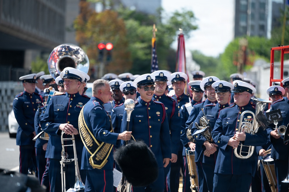 A military marching band is assembled outdoors, likely preparing for or participating in a parade. Members are dressed in formal dark navy blue uniforms with gold buttons, white peaked caps, and red trim, carrying brass instruments. An American flag and what appears to be a unit color guard flag are visible in the background.