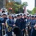 A military marching band is assembled outdoors, likely preparing for or participating in a parade. Members are dressed in formal dark navy blue uniforms with gold buttons, white peaked caps, and red trim, carrying brass instruments. An American flag and what appears to be a unit color guard flag are visible in the background.