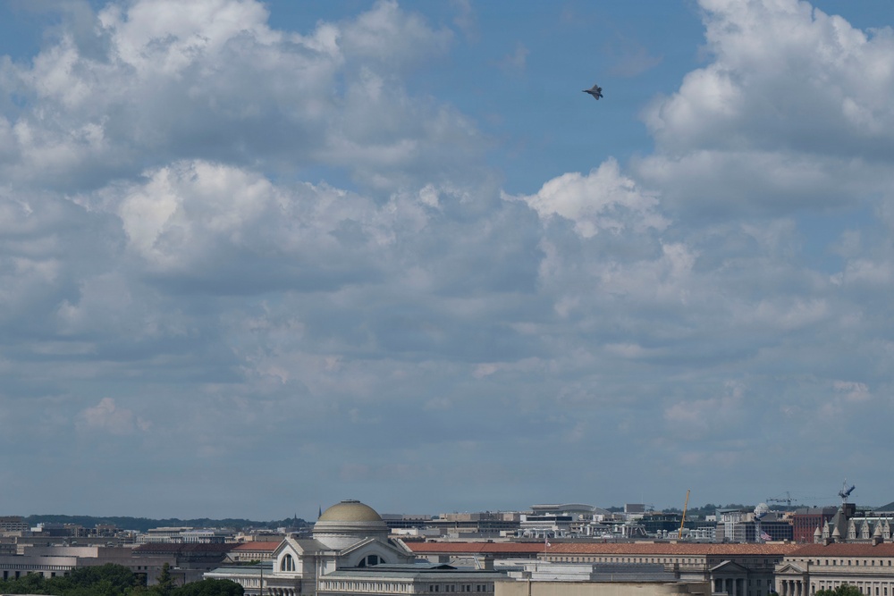 An aerial view of Washington D.C.'s skyline featuring neoclassical government buildings and museums beneath a partly cloudy blue sky. A military jet aircraft is visible flying at altitude in the upper portion of the frame. The scene appears to be taken from an elevated vantage point, possibly from a rooftop or elevated structure.