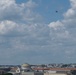 An aerial view of Washington D.C.'s skyline featuring neoclassical government buildings and museums beneath a partly cloudy blue sky. A military jet aircraft is visible flying at altitude in the upper portion of the frame. The scene appears to be taken from an elevated vantage point, possibly from a rooftop or elevated structure.