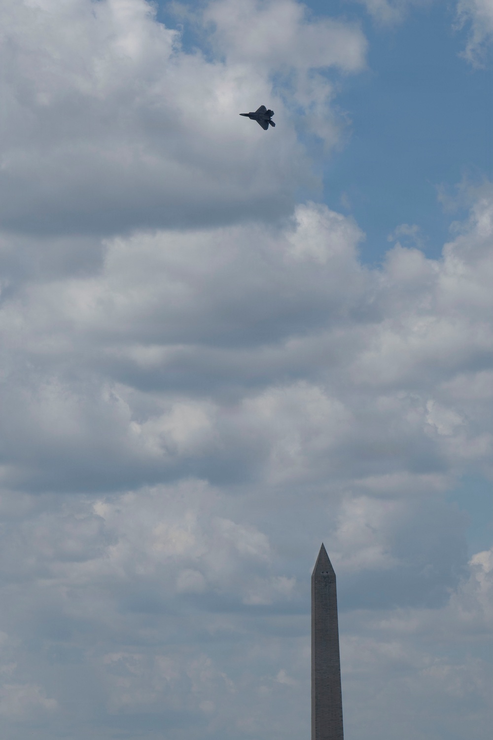 A military jet aircraft, consistent with an F-22 Raptor silhouette, flies above the Washington Monument in Washington, D.C. The image is taken from a low angle looking upward, capturing both the iconic obelisk monument and the aircraft against a partly cloudy blue sky. This appears to be a flyover event, likely associated with a national celebration or military demonstration.