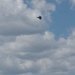 A military jet aircraft, consistent with an F-22 Raptor silhouette, flies above the Washington Monument in Washington, D.C. The image is taken from a low angle looking upward, capturing both the iconic obelisk monument and the aircraft against a partly cloudy blue sky. This appears to be a flyover event, likely associated with a national celebration or military demonstration.