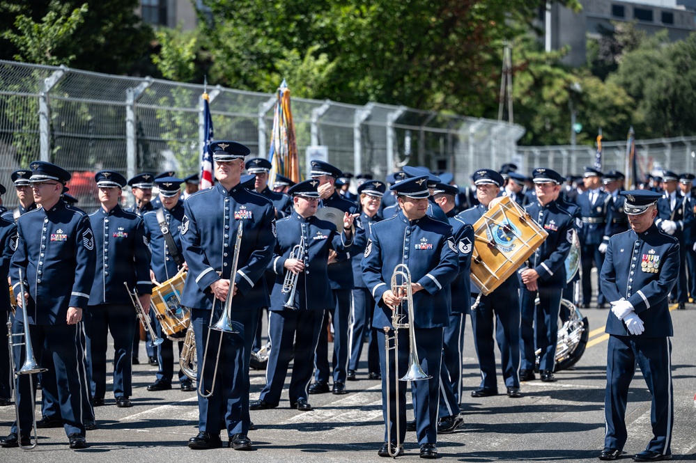 A United States Air Force military band is assembled on a paved surface, likely preparing for or participating in a parade or ceremonial event. Personnel are dressed in formal dark blue Air Force dress uniforms with service ribbons and rank insignia, carrying various musical instruments. An American flag and what appears to be a unit guidon are visible in the background.