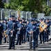 A United States Air Force military band is assembled on a paved surface, likely preparing for or participating in a parade or ceremonial event. Personnel are dressed in formal dark blue Air Force dress uniforms with service ribbons and rank insignia, carrying various musical instruments. An American flag and what appears to be a unit guidon are visible in the background.