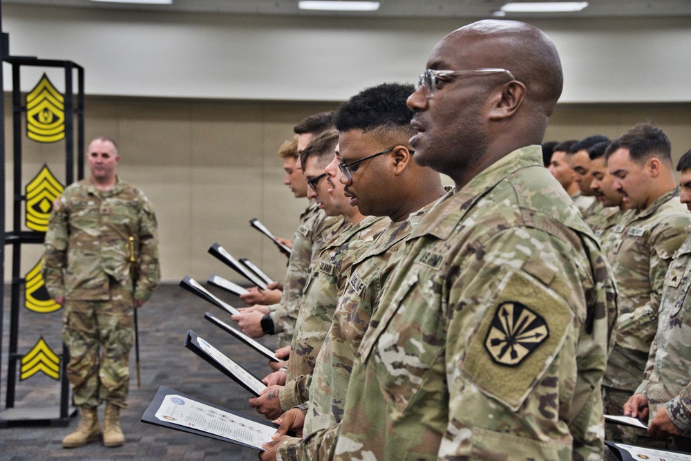 A formal U.S. Army military ceremony is taking place indoors, with a line of soldiers in OCP (Operational Camouflage Pattern) uniforms holding open document folders or certificates. A senior non-commissioned officer stands at the front facing the formation, with large Sergeant Major rank insignia displayed on a backdrop behind him. The setting appears to be a promotion or graduation ceremony.