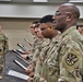 A formal U.S. Army military ceremony is taking place indoors, with a line of soldiers in OCP (Operational Camouflage Pattern) uniforms holding open document folders or certificates. A senior non-commissioned officer stands at the front facing the formation, with large Sergeant Major rank insignia displayed on a backdrop behind him. The setting appears to be a promotion or graduation ceremony.