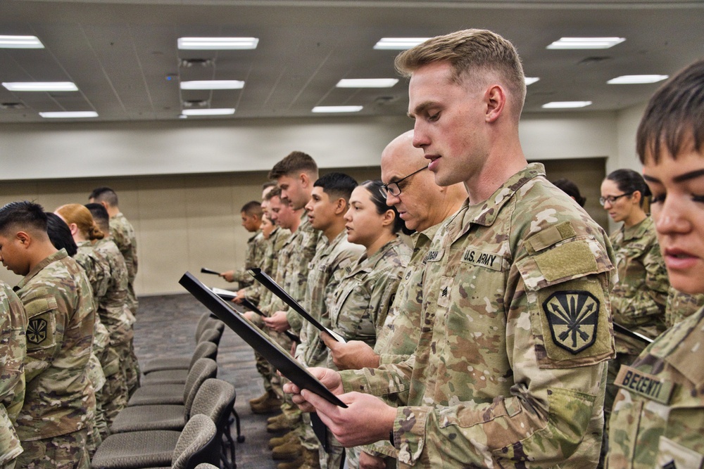 A group of U.S. Army soldiers in Operational Camouflage Pattern (OCP) uniforms are standing in rows inside an indoor facility, reading from black-framed documents or certificates. The setting appears to be a formal military ceremony, likely a promotion, award, or oath-of-office event. Multiple soldiers of varying ranks and backgrounds are visible, all focused on the documents they are holding.